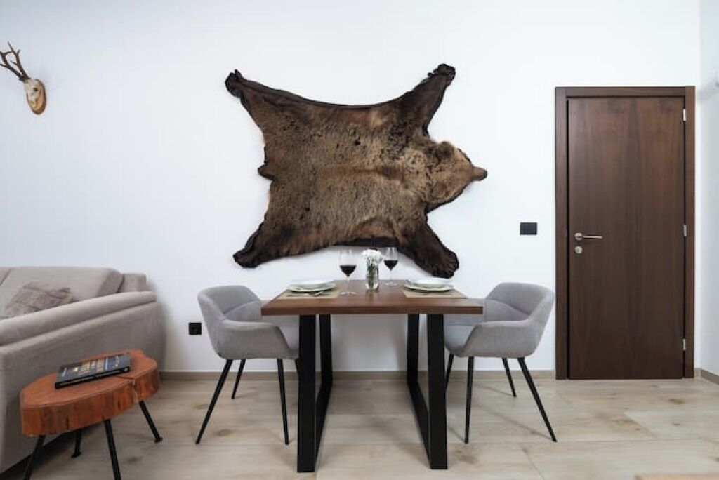 A dining area with a wooden table, two grey chairs, a small round wooden side table, and a mounted animal hide on the wall.