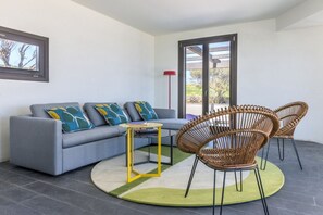 Un salon moderne avec un canapé gris, des fauteuils en osier, une petite table jaune et un tapis vert.