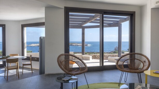 Una habitación moderna con ventanas amplias, sillas de mimbre, una mesa redonda y vistas al mar.
