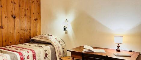 Una habitación con pared de madera, una cama con ropa de cama estampada, un escritorio con una lámpara y una silla.
