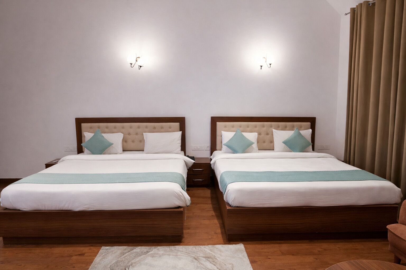 Quarto de hotel com duas camas, cabeceira de madeira, roupa de cama branca, tapete bege e cortina marrom.