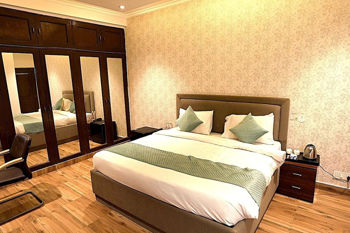 A hotel room with a large bed, a mirror cabinet, a chair, and a bedside table with a kettle.