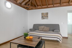 A room with a bed, a table with fruit, a plant, and a painting on the wall.