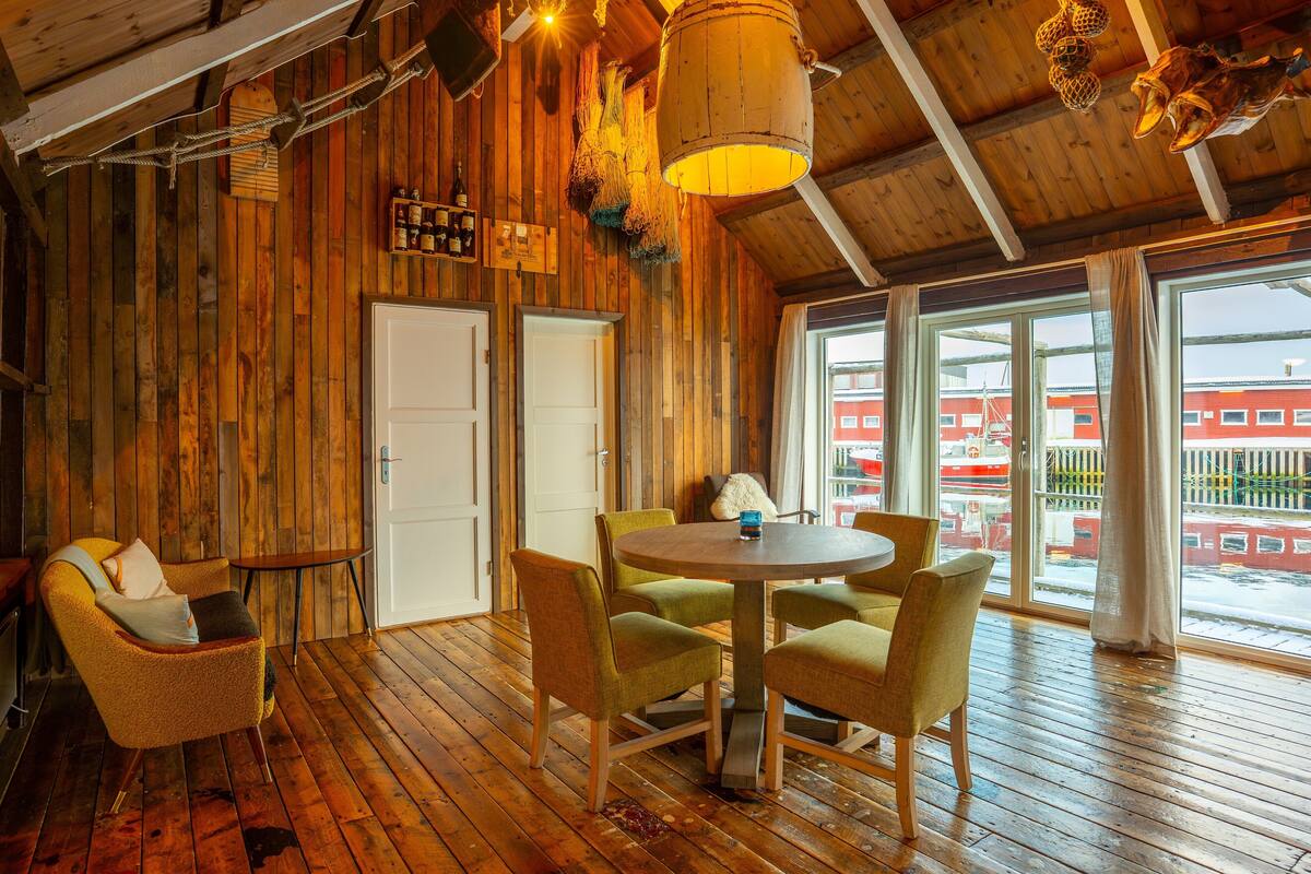 A wooden interior with a round table and chairs, a white door, and large windows with a view of a red building outside.