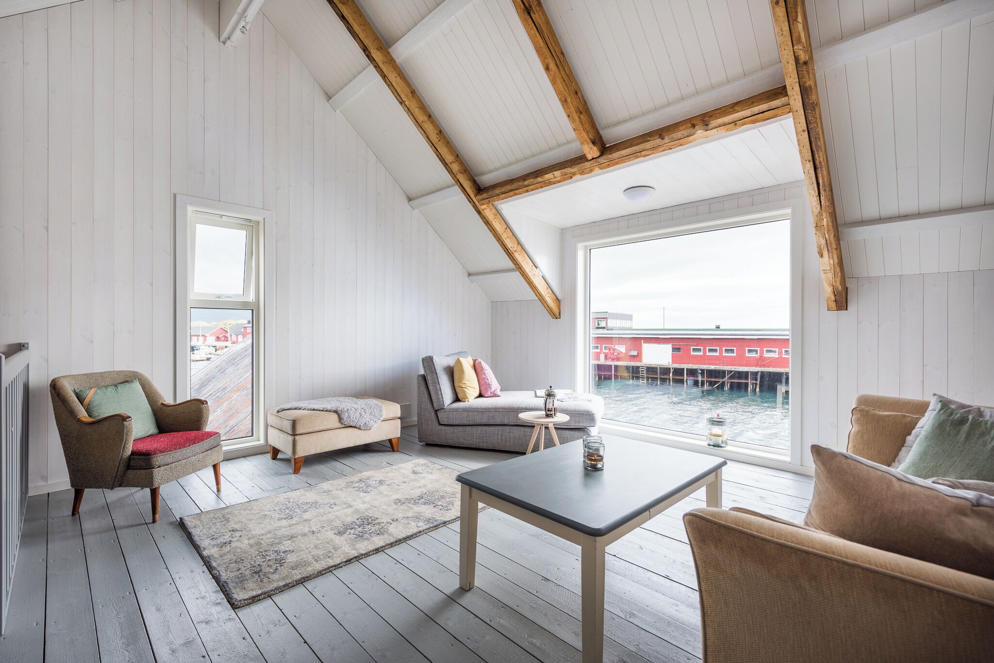 Ein geräumiges Schlafzimmer im Loft-Stil mit hohen Decken, Holzbalken, einem großen Fenster mit Blick auf ein Gewässer, einer gemütlichen Sitzecke mit einer Couch und Sesseln sowie einem kleinen Tisch.