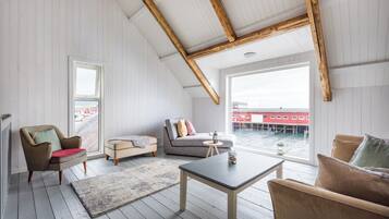 Amplio dormitorio tipo loft con techo alto, vigas de madera, un ventanal grande con vistas a un cuerpo de agua, una zona de estar acogedora con sofá y sillones, y una mesita.