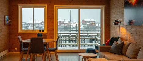 A cozy room with wooden walls, a dining table with chairs, a sofa, and large windows overlooking a snowy dock area.