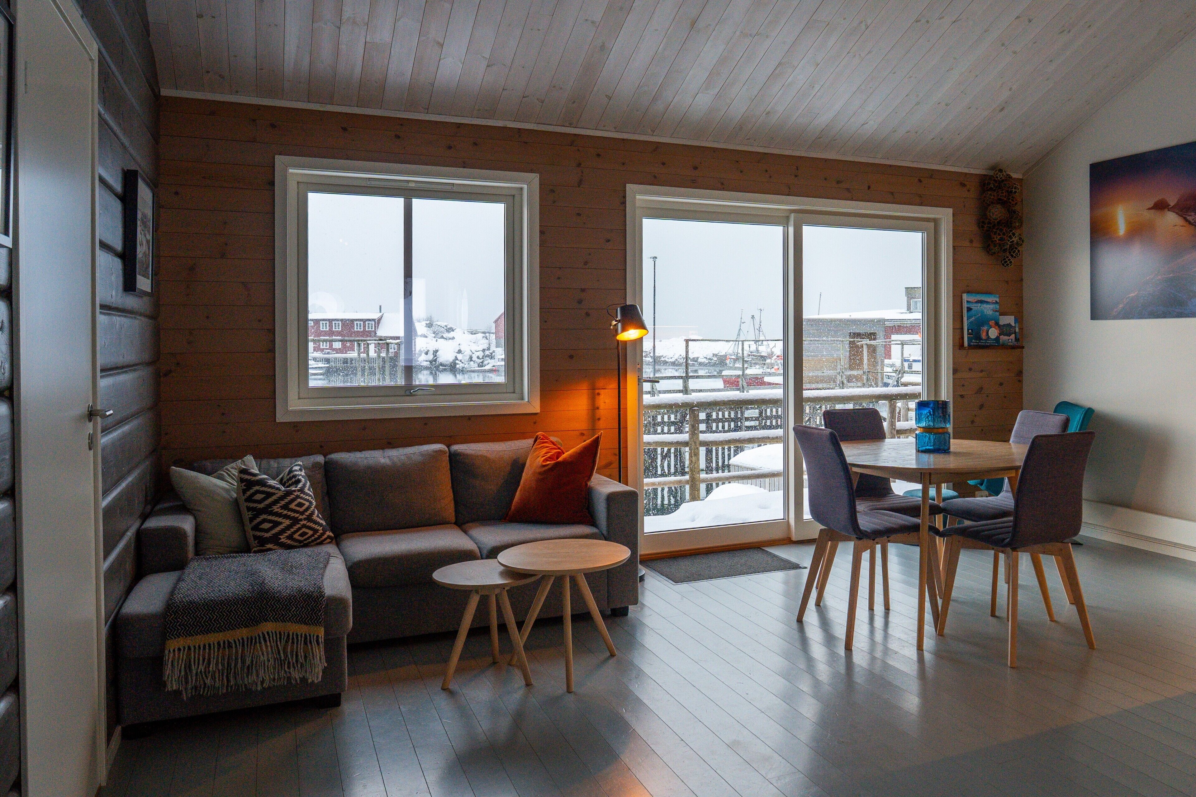 Een gezellige woonkamer met een bank, twee kleine tafels en een eettafel met stoelen. Grote ramen bieden uitzicht op een besneeerd landschap.