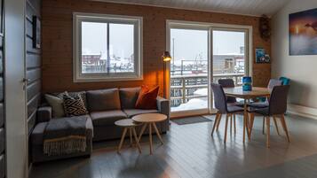 A cozy living room with a sofa, two small tables, and a dining table with chairs. Large windows offer a view of a snowy landscape.