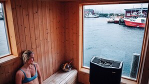 Une personne vêtue d’un maillot de bain bleu est assise sur un banc en bois à l’intérieur d’un chalet, d’où l’on peut admirer des bateaux sur l’eau.