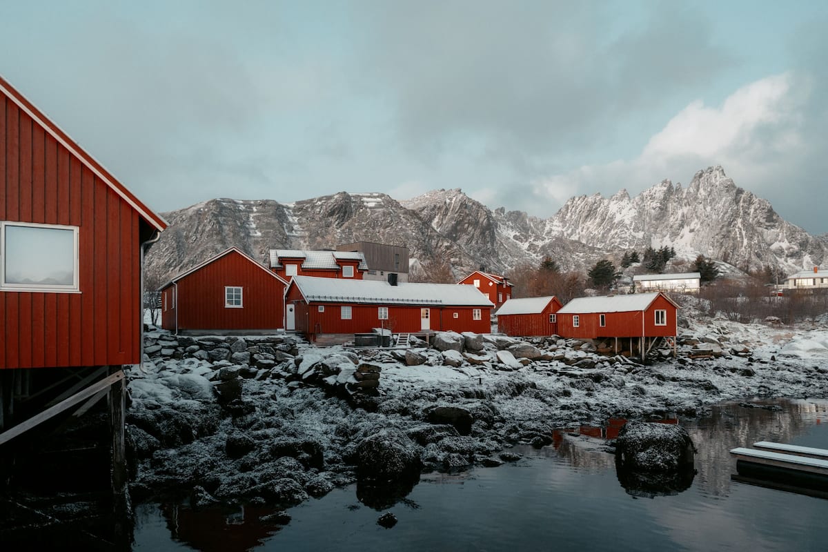 A row of red wooden buildings by a frozen lake with snow-covered mountains in the background.