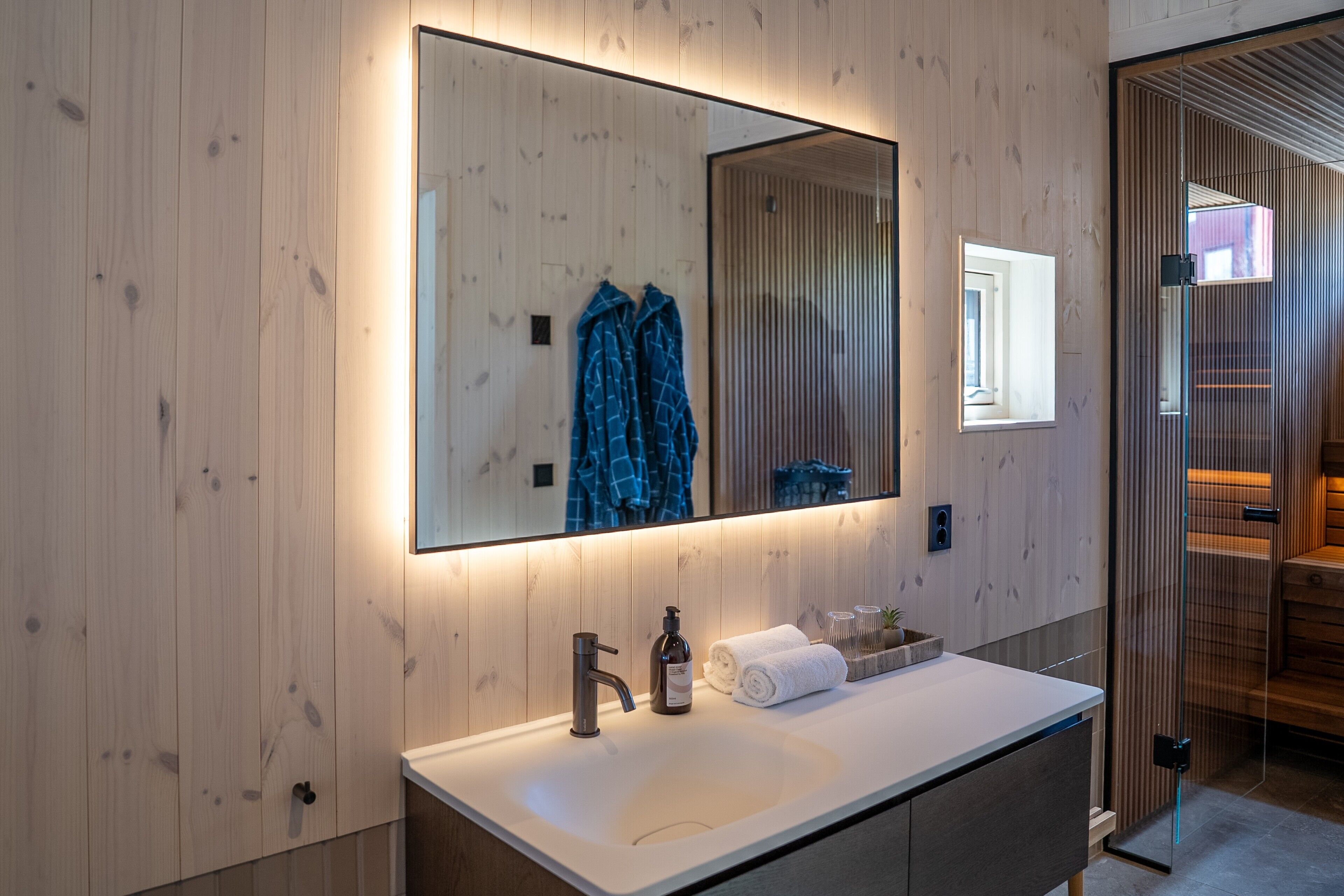 A modern bathroom with a wooden wall, a large mirror, a sink, a faucet, and a glass partition.
