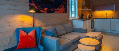 Una sala moderna con sofá, sillón y reposapiés, un cuadro en la pared y una zona de cocina con armarios y ventanal.