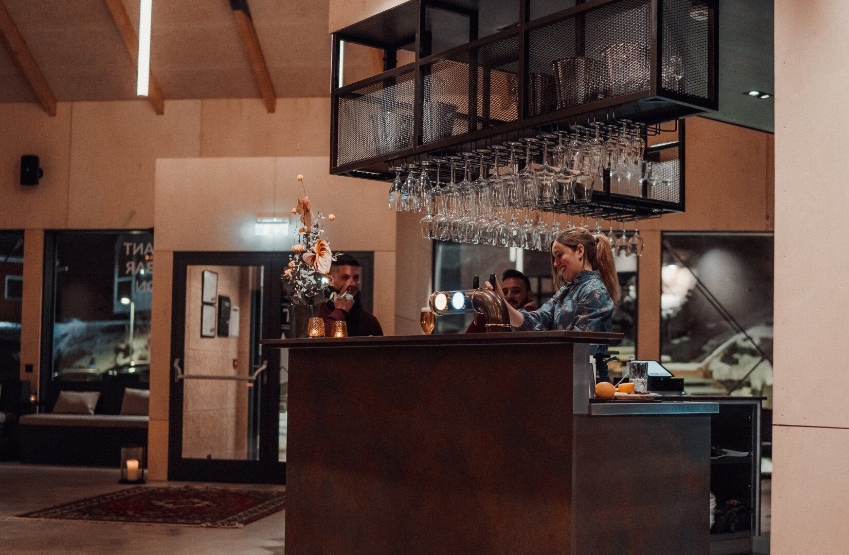Una zona de bar con un barman y un cliente, una lámpara colgante y una estantería de vinos en la pared.