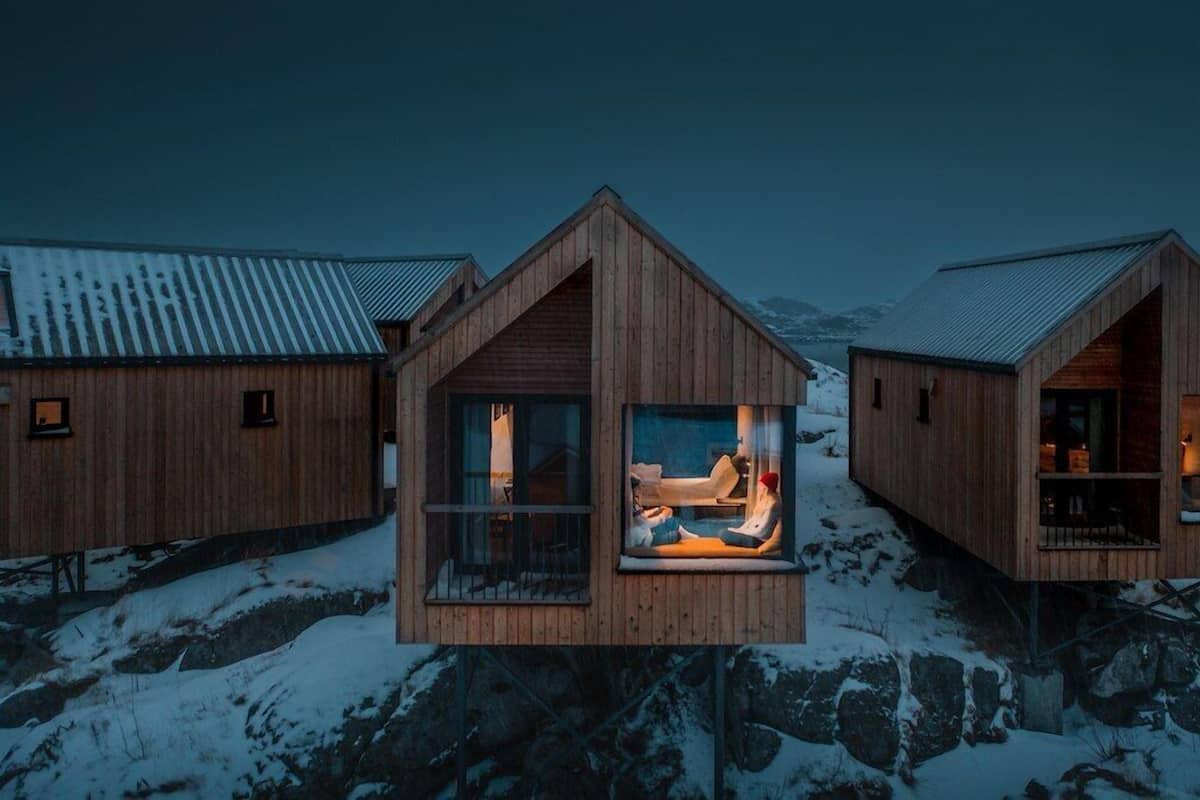A wooden cabin with a large window, a metal roof, and a snowy landscape in the background.