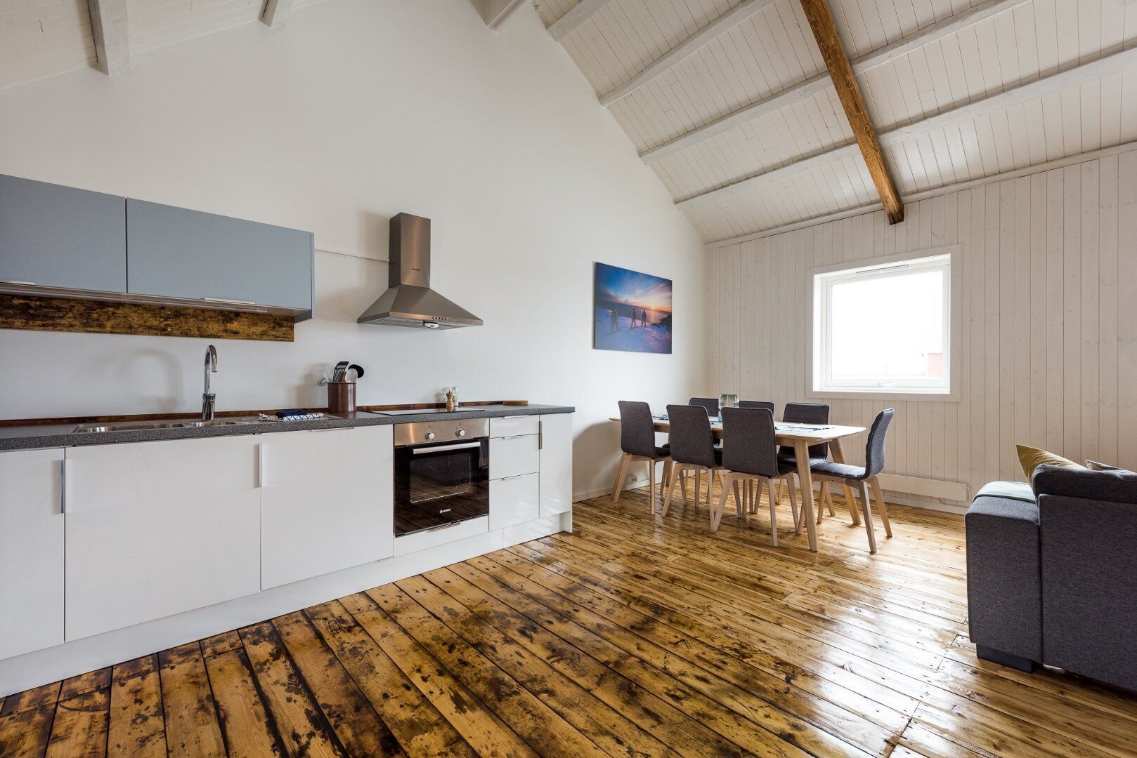 A modern kitchen with white cabinets, a stainless steel sink, and a built-in oven. There is a dining area with a table and chairs, and a large window providing natural light.