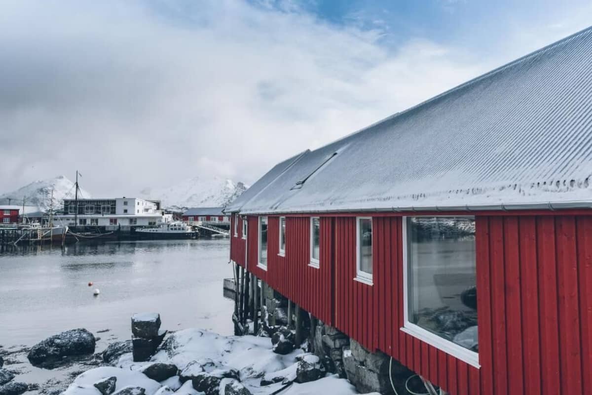 A red building with a sloped roof, situated by a calm body of water, with snow-covered rocks in the foreground and mountains in the background.