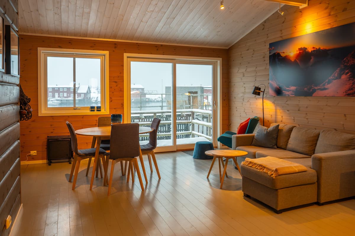 A cozy living room with a wooden floor, a sofa, a dining table with chairs, and a large window with a view of buildings.