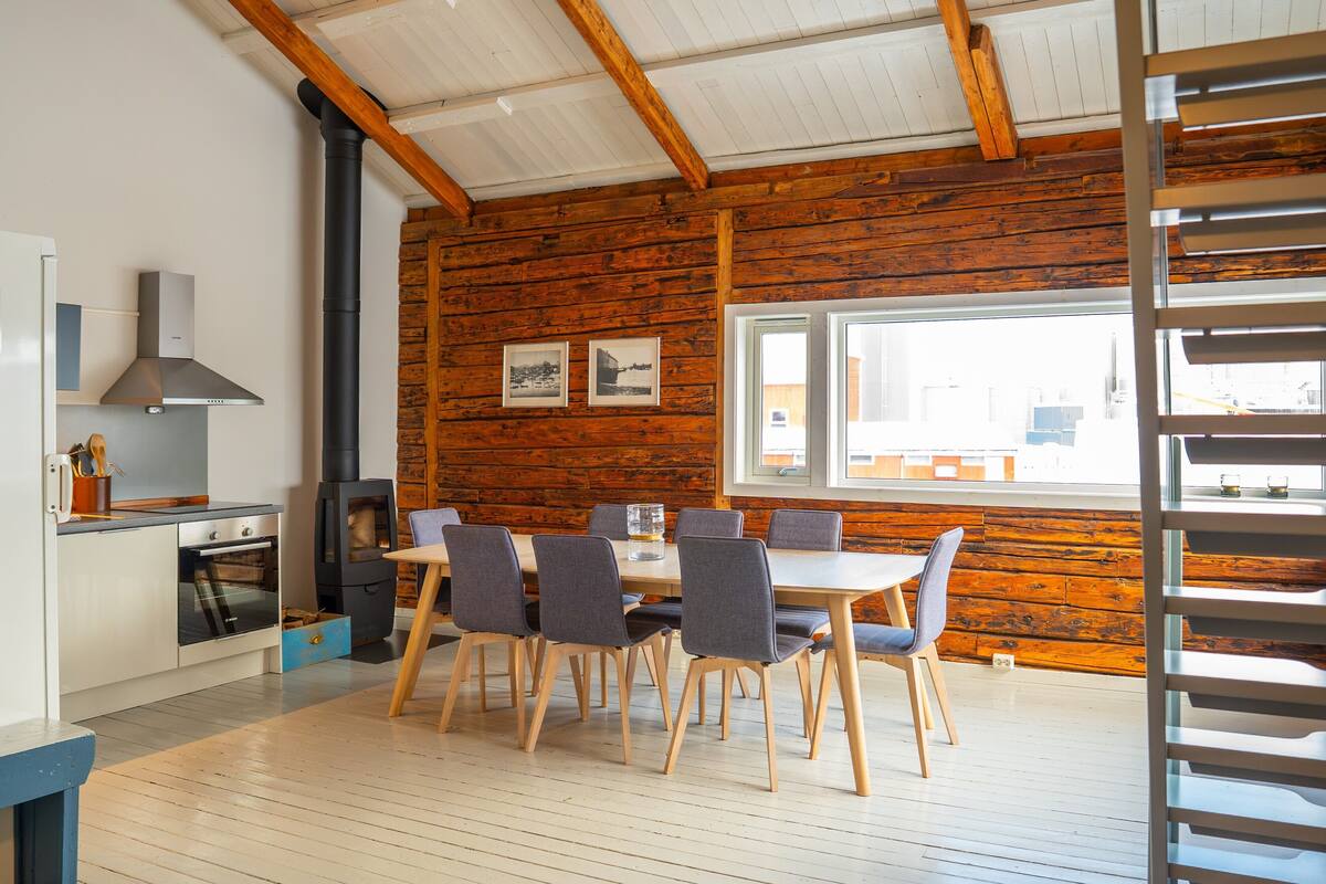 A modern kitchen with wooden walls, a dining area with six chairs, and a large window.