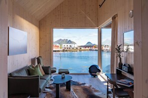 A modern interior with a sofa, coffee table, and a view of a waterfront with buildings and mountains. - Hattvika Lodge (Vestvagoy)