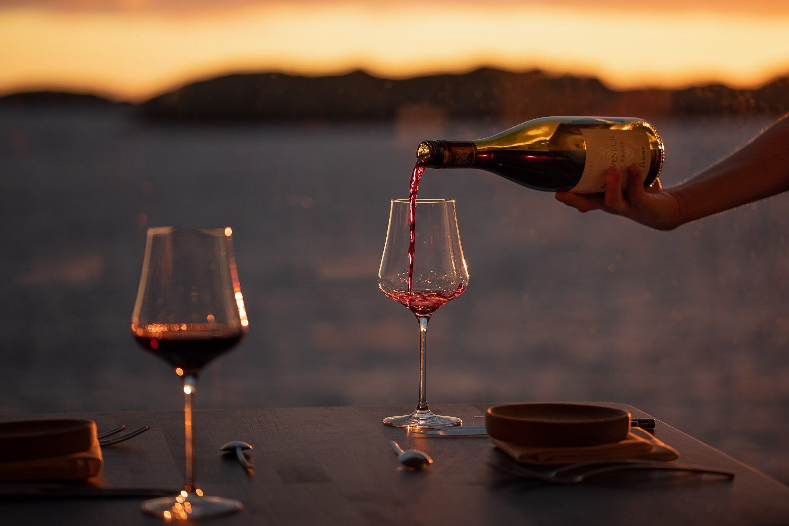 A bottle of wine being poured into a glass at a table with another glass and a bowl, set against a sunset backdrop.