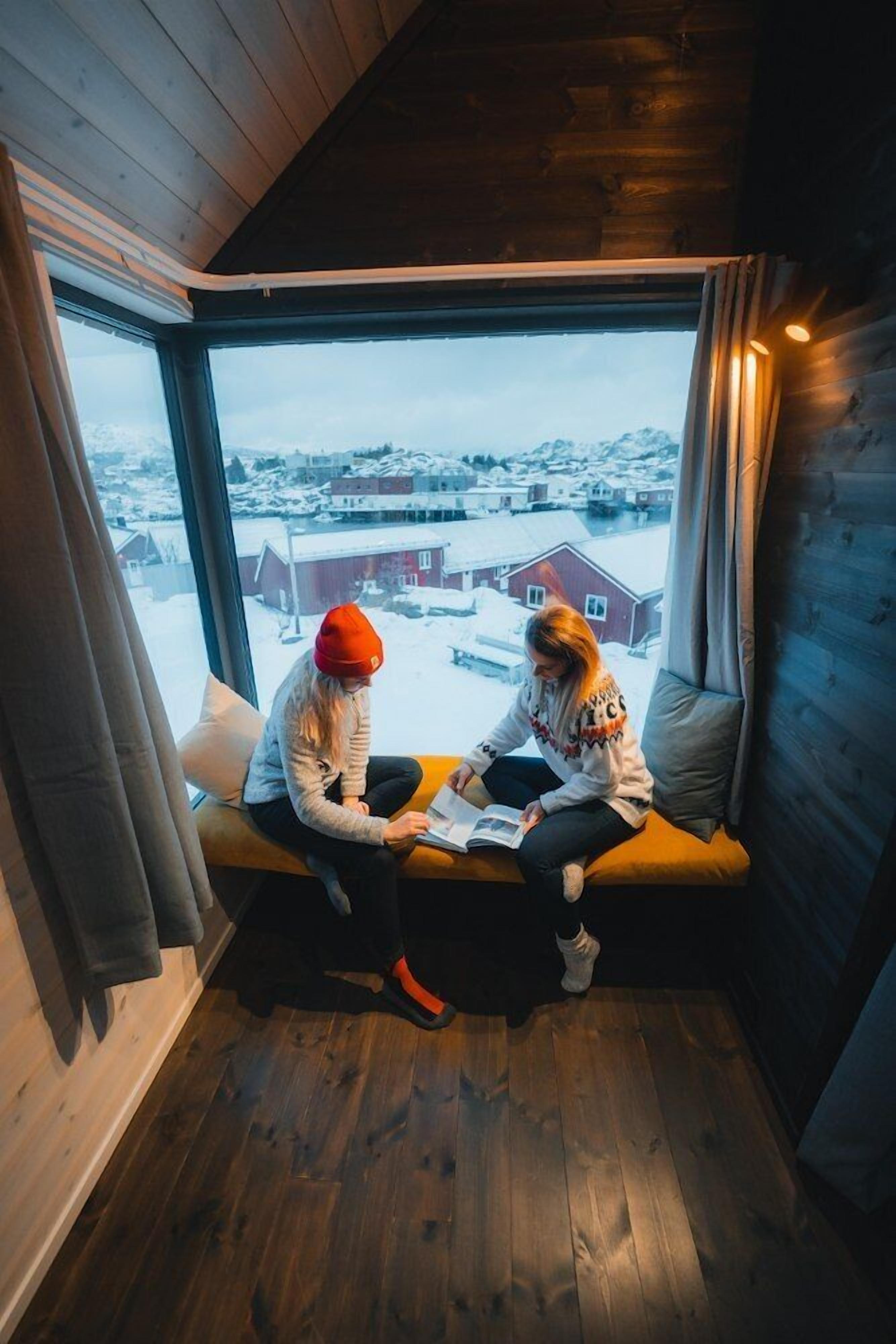 Deux personnes sont assises sur un banc en bois, près d’une grande fenêtre qui donne sur un paysage enneigé.