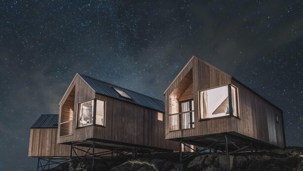Deux cabanes en bois, surélevées sur des piliers, dotées de grandes fenêtres, se dressent sur fond de ciel étoilé.