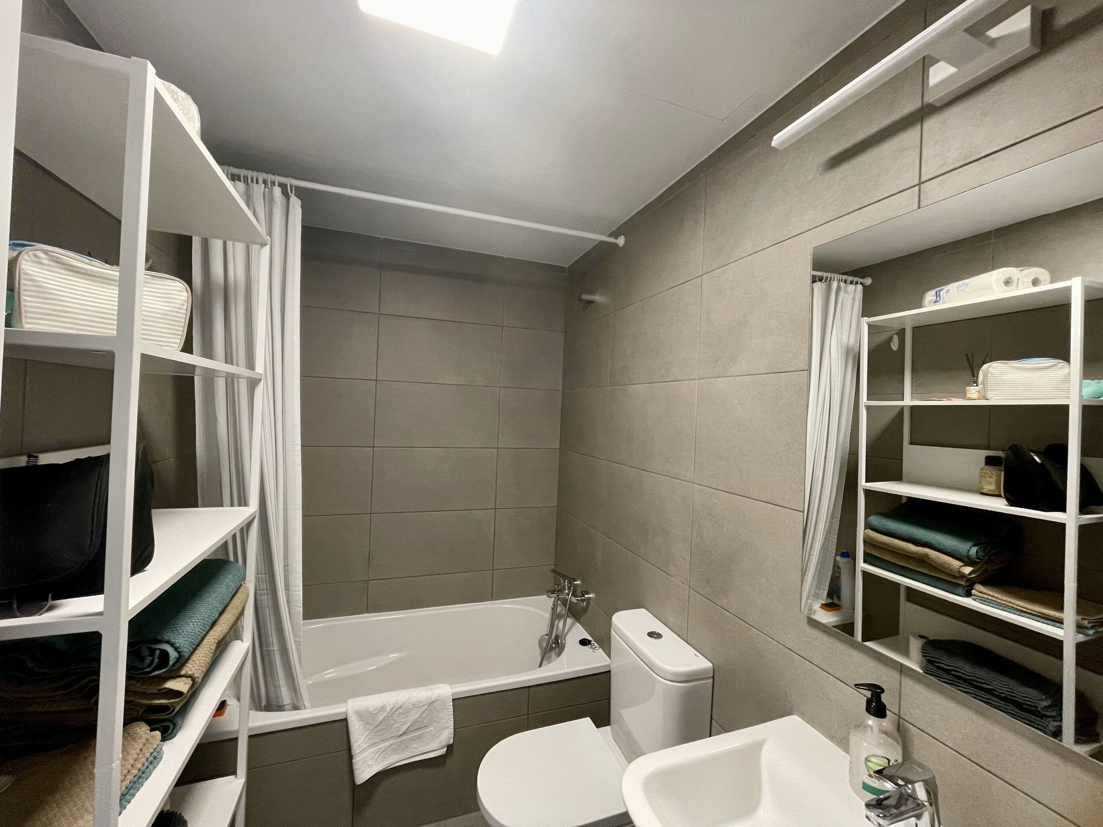 A modern bathroom with a bathtub, toilet, and shelves with folded towels and toiletries.