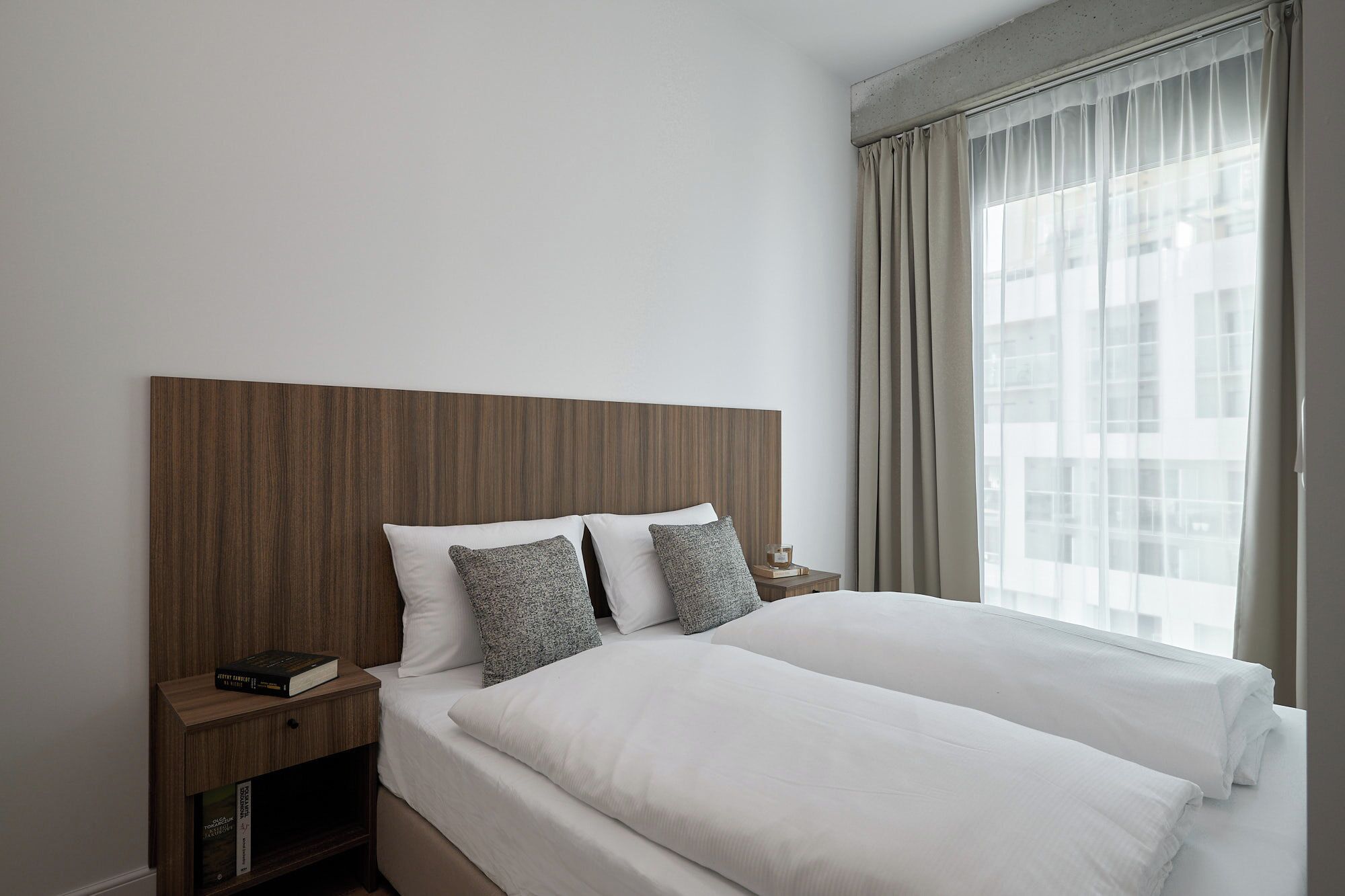 Een moderne hotelkamer met een groot bed, een houten hoofdbord, een nachtkastje met een boek en uitzicht op gebouwen door dunne gordijnen.