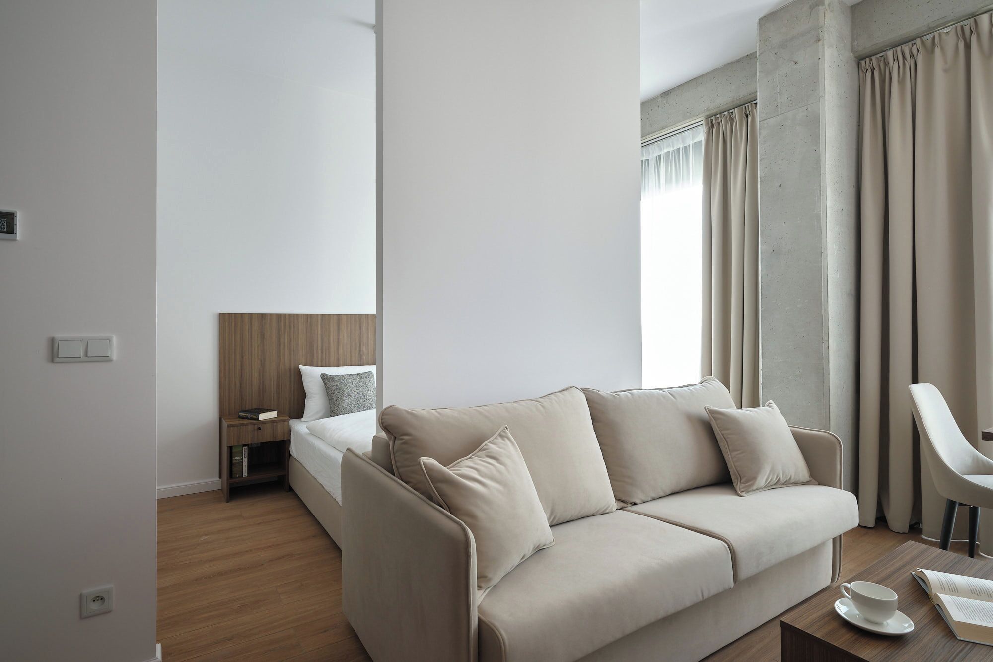 Una habitación de hotel moderna con sofá, mesita de noche, silla y ventana con cortinas.