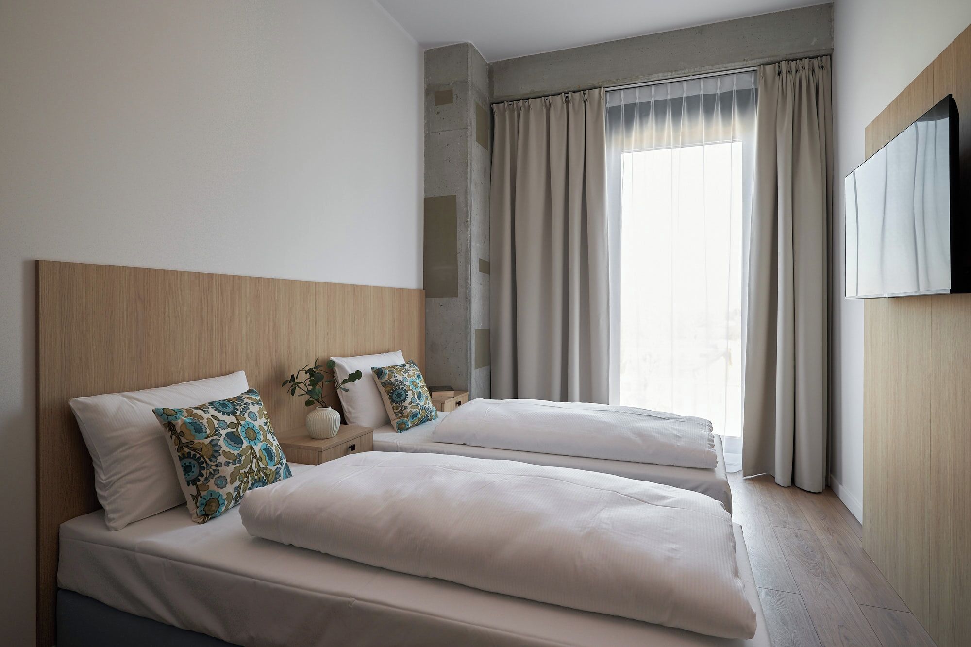 Une chambre d’hôtel avec une tête de lit en bois, deux lits avec du linge de lit blanc, une grande fenêtre avec des rideaux et un téléviseur à écran plat fixé au mur.