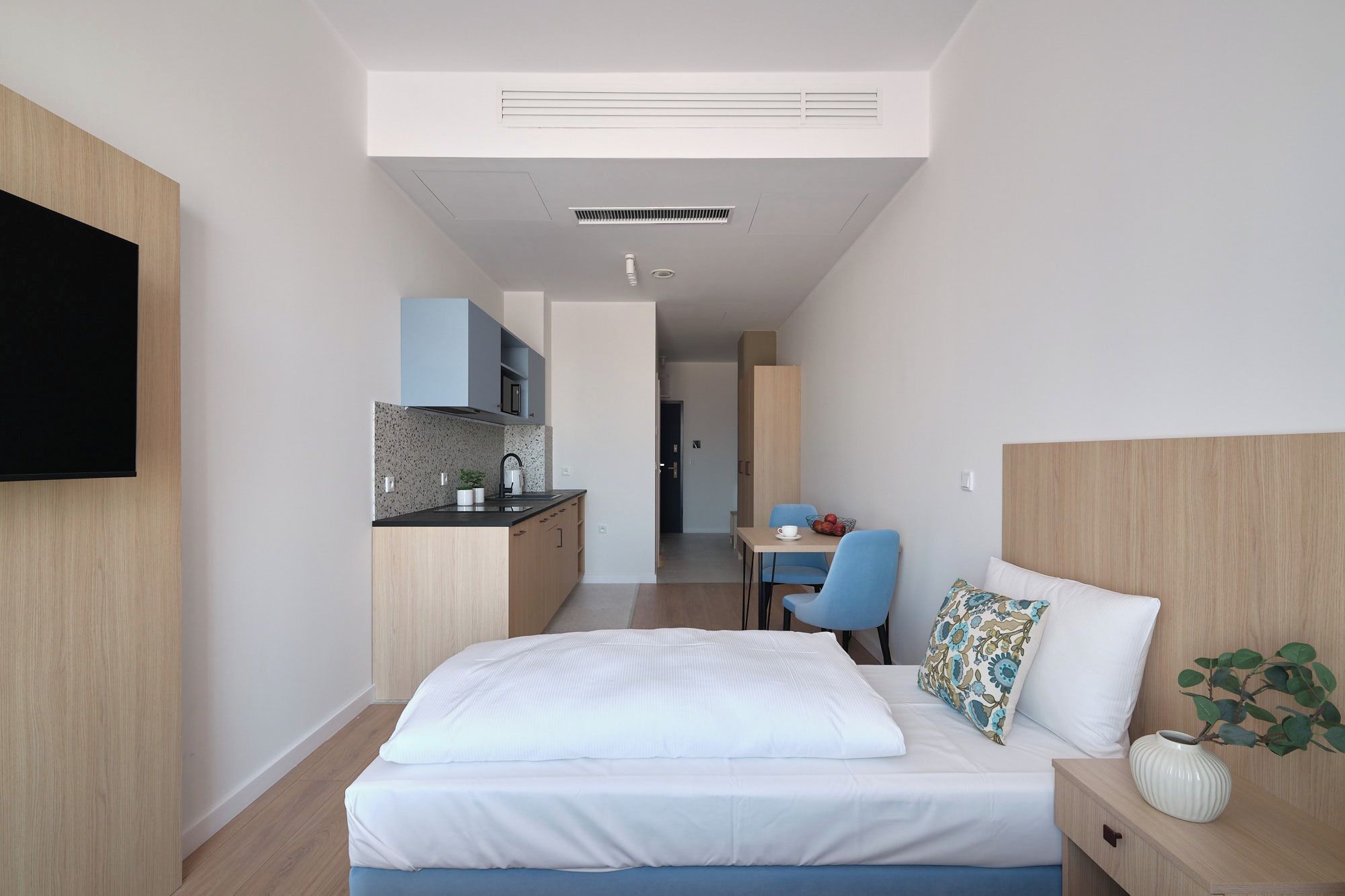 A modern hotel room with a bed, bedside table, a small desk with a chair, a kitchenette, and a TV mounted on the wall.