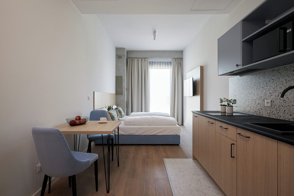 Une chambre d’hôtel de taille réduite, comprenant un lit, une petite table à manger avec des chaises, un coin cuisine et une fenêtre avec des rideaux.