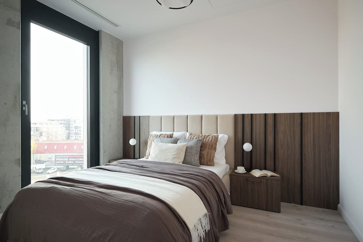 Une chambre moderne avec une grande fenêtre, un lit agrémenté de plusieurs oreillers, une table de chevet avec une tasse et un livre, et une tête de lit en bois.