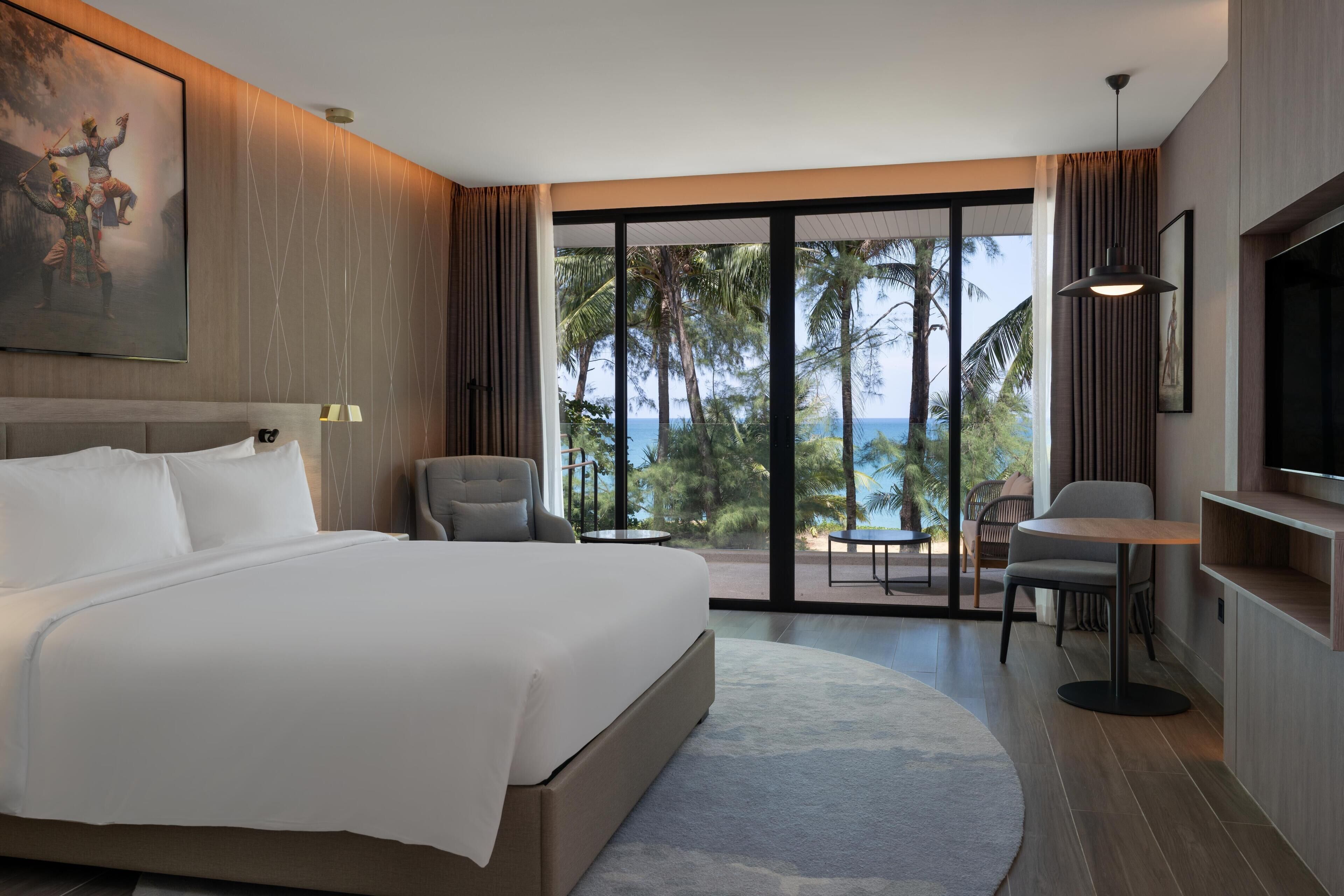 Camera d'albergo moderna con un letto grande, una televisione, una scrivania e un balcone vista palme e oceano.