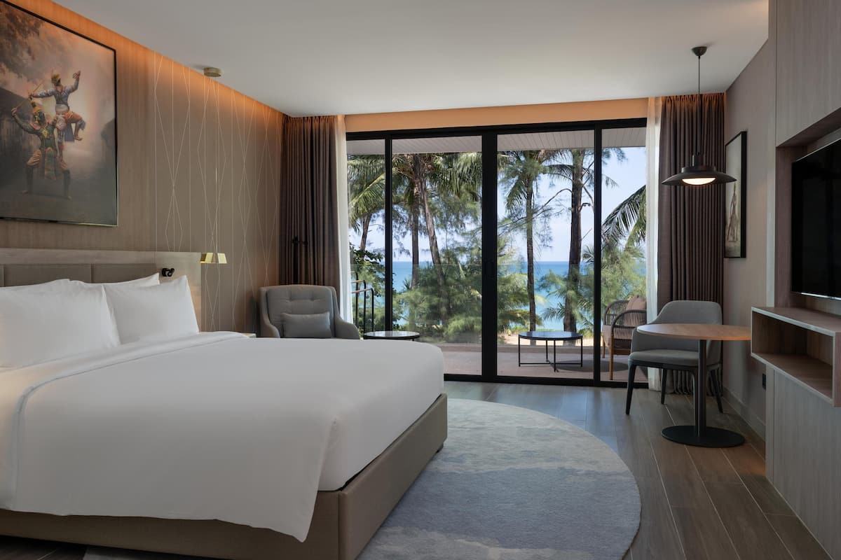 Una habitación de hotel moderna con una cama grande, televisión, escritorio y balcón con vistas a palmeras y al océano.