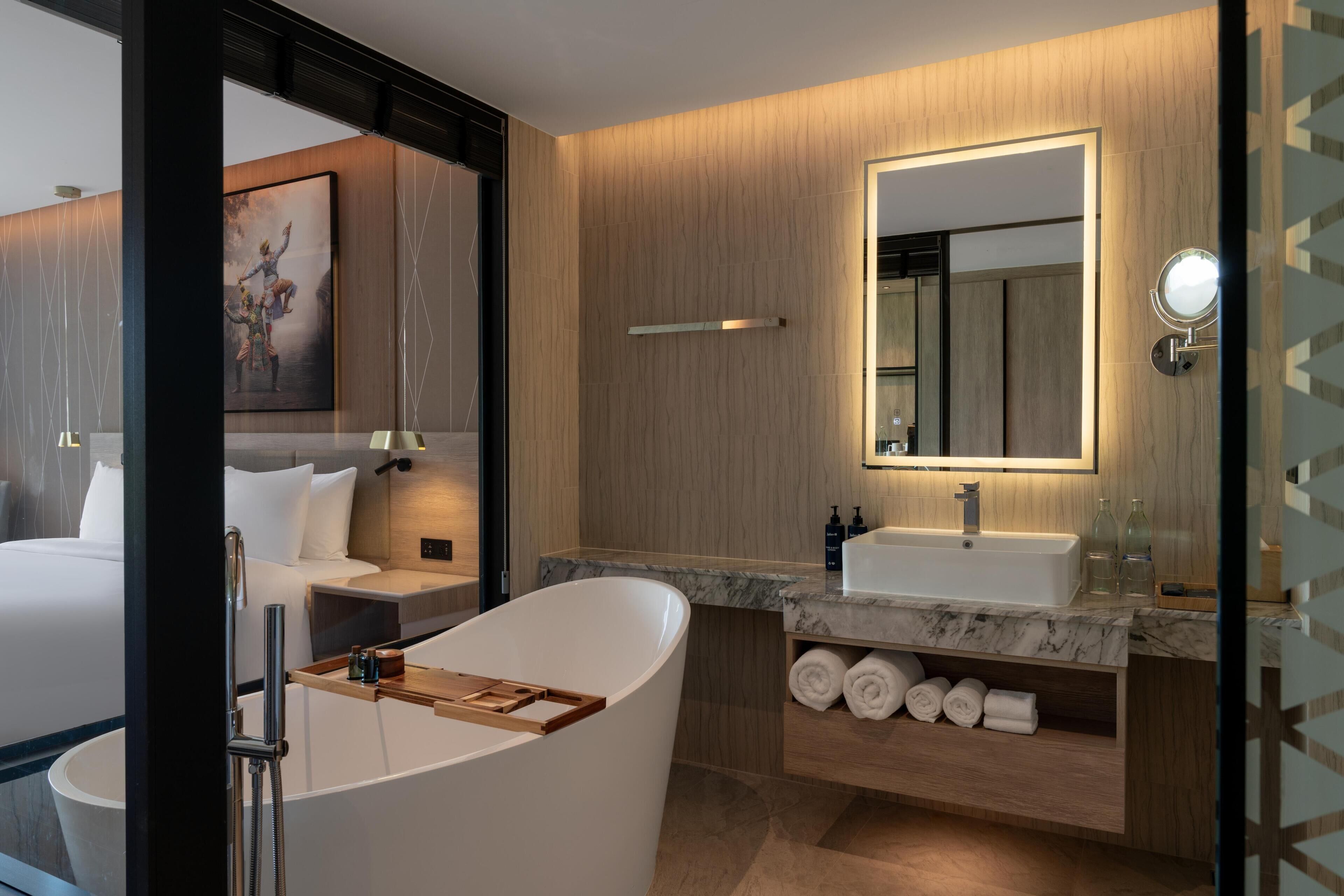 A modern bathroom with a bathtub, a vanity with a mirror, and a bed visible through an open door.