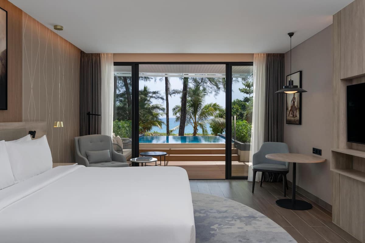 Habitación de hotel con una cama grande, un área de descanso con una mesa, un televisor y vistas a una piscina y palmeras.