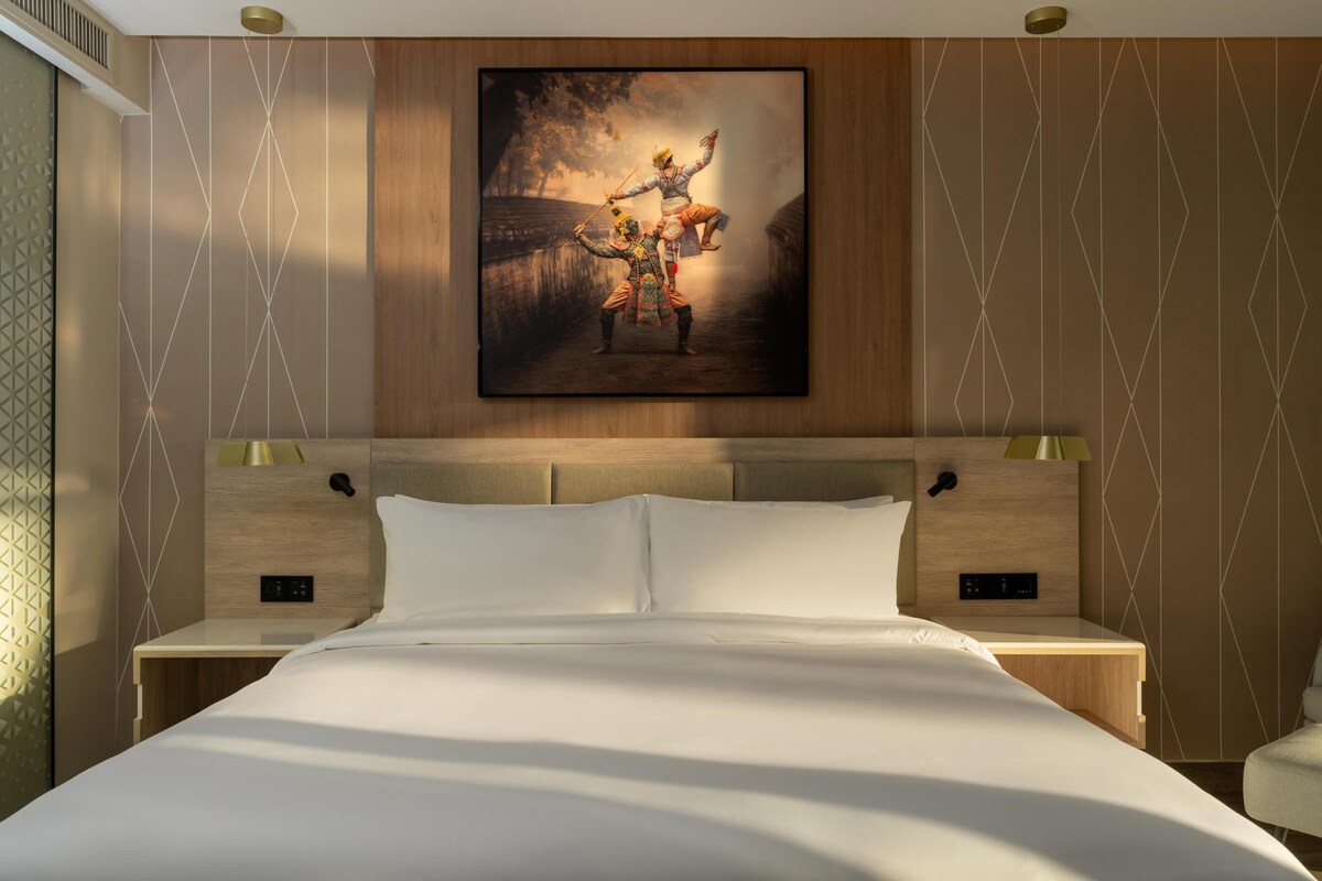 Une chambre d’hôtel avec un grand lit, deux tables de chevet, une tête de lit dotée d’une lampe intégrée et un tableau encadré au mur.