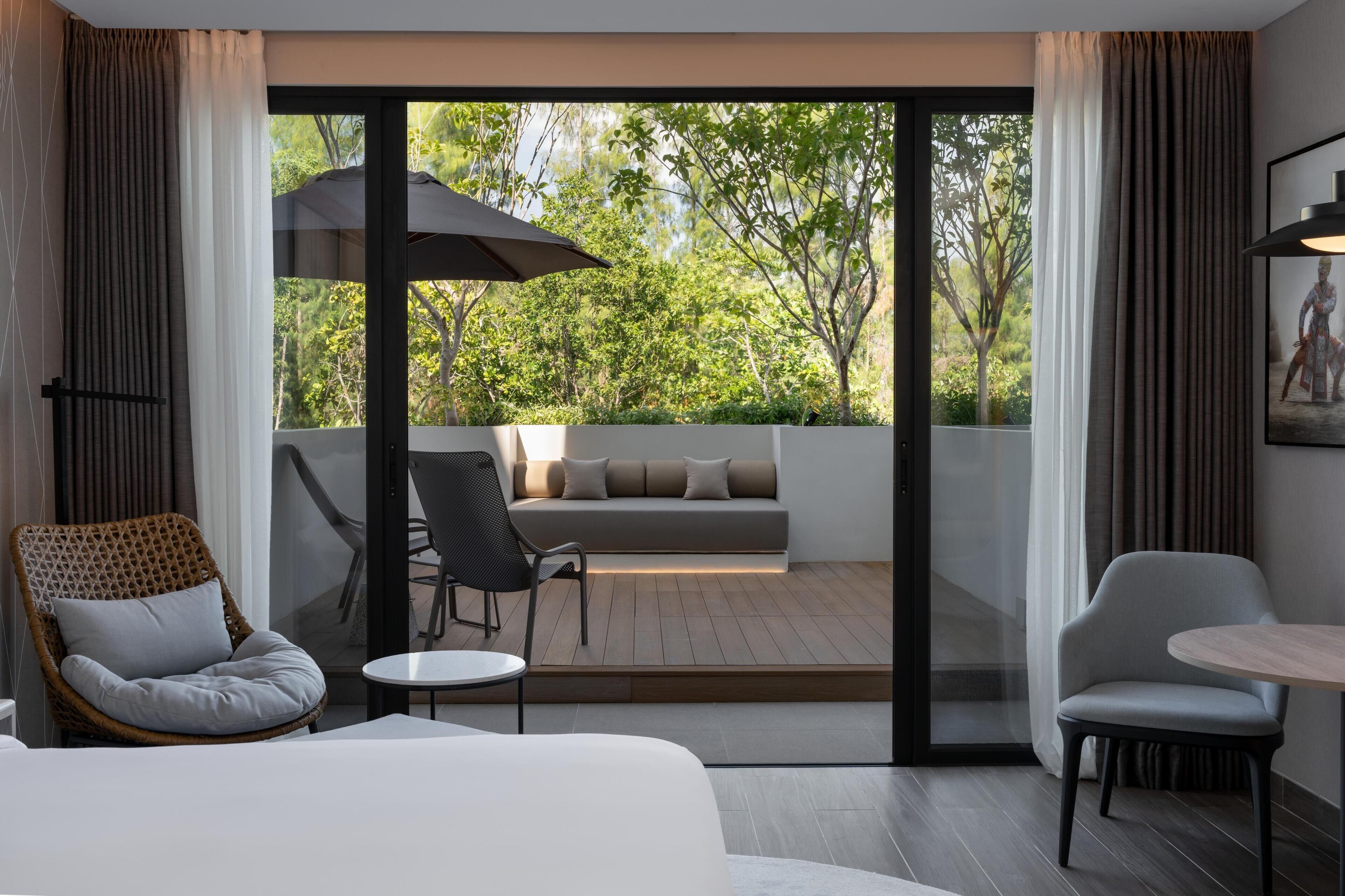 A modern hotel room with a large sliding glass door leading to an outdoor seating area, a bed, a small table, and a wicker chair.