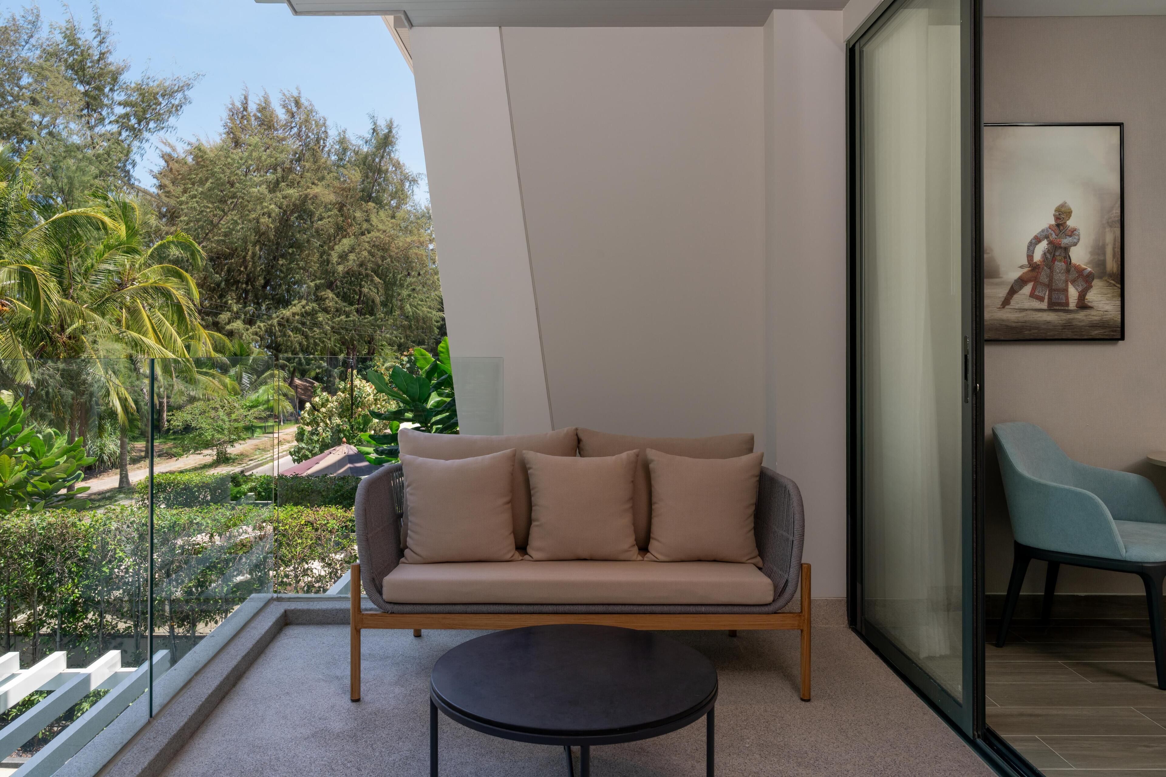 A modern living room with a sofa, a round table, and a glass door leading to a balcony with a view of a garden and trees.