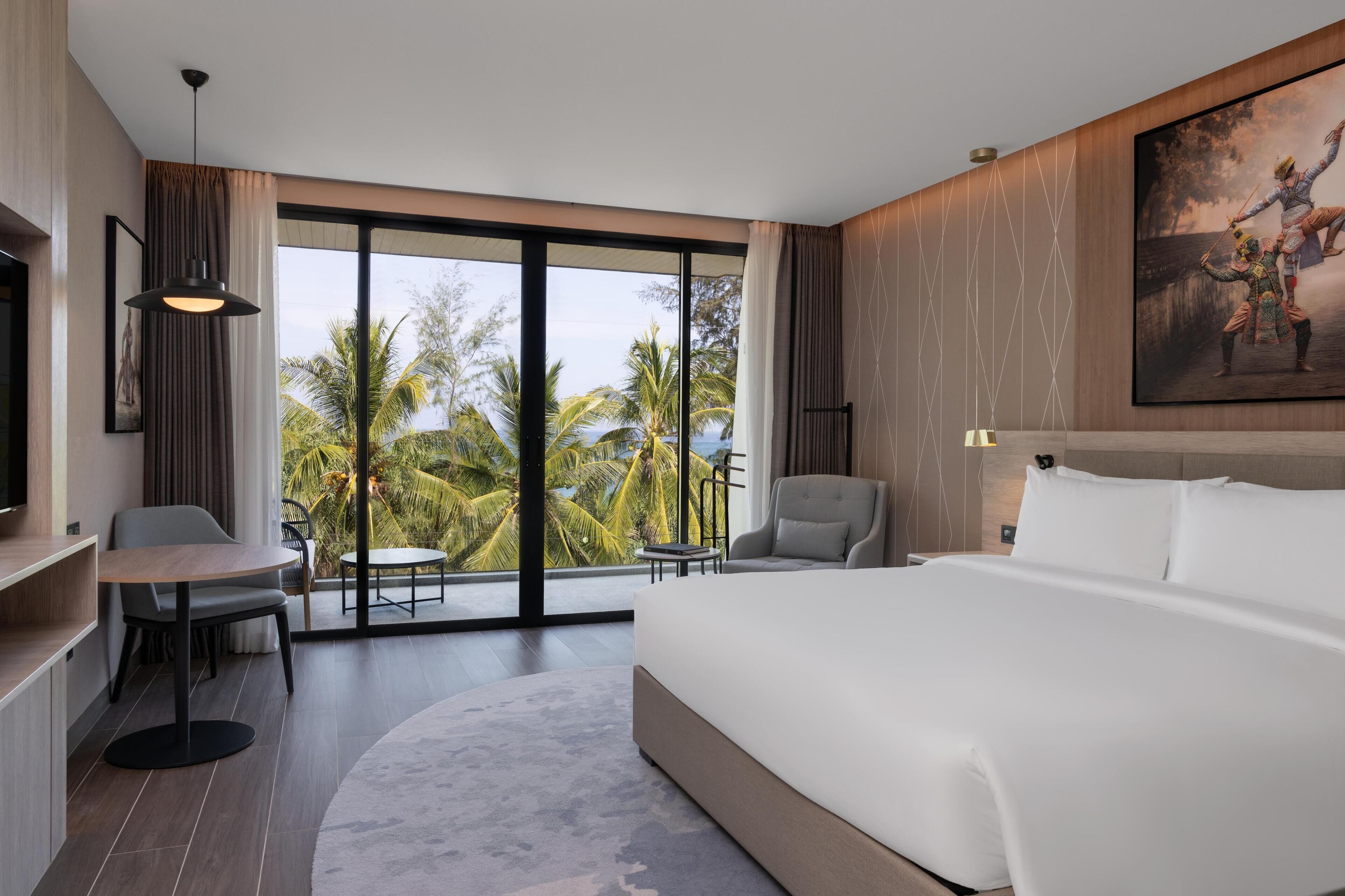A modern hotel room with a large bed, a desk, a chair, and a view of palm trees through large windows.