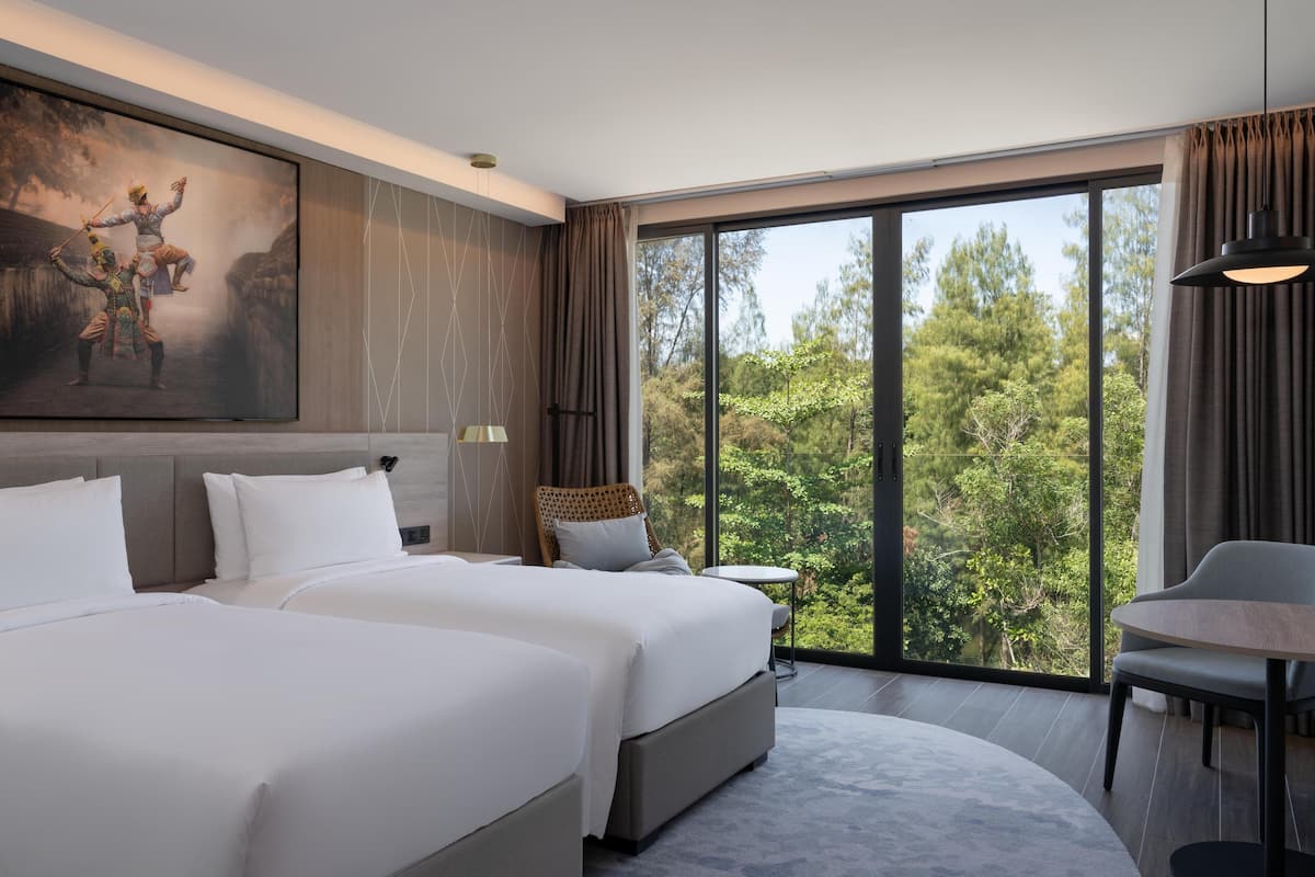 Habitación de hotel con dos camas, un ventanal con vistas a árboles, un rincón de comedor y un cuadro en la pared.