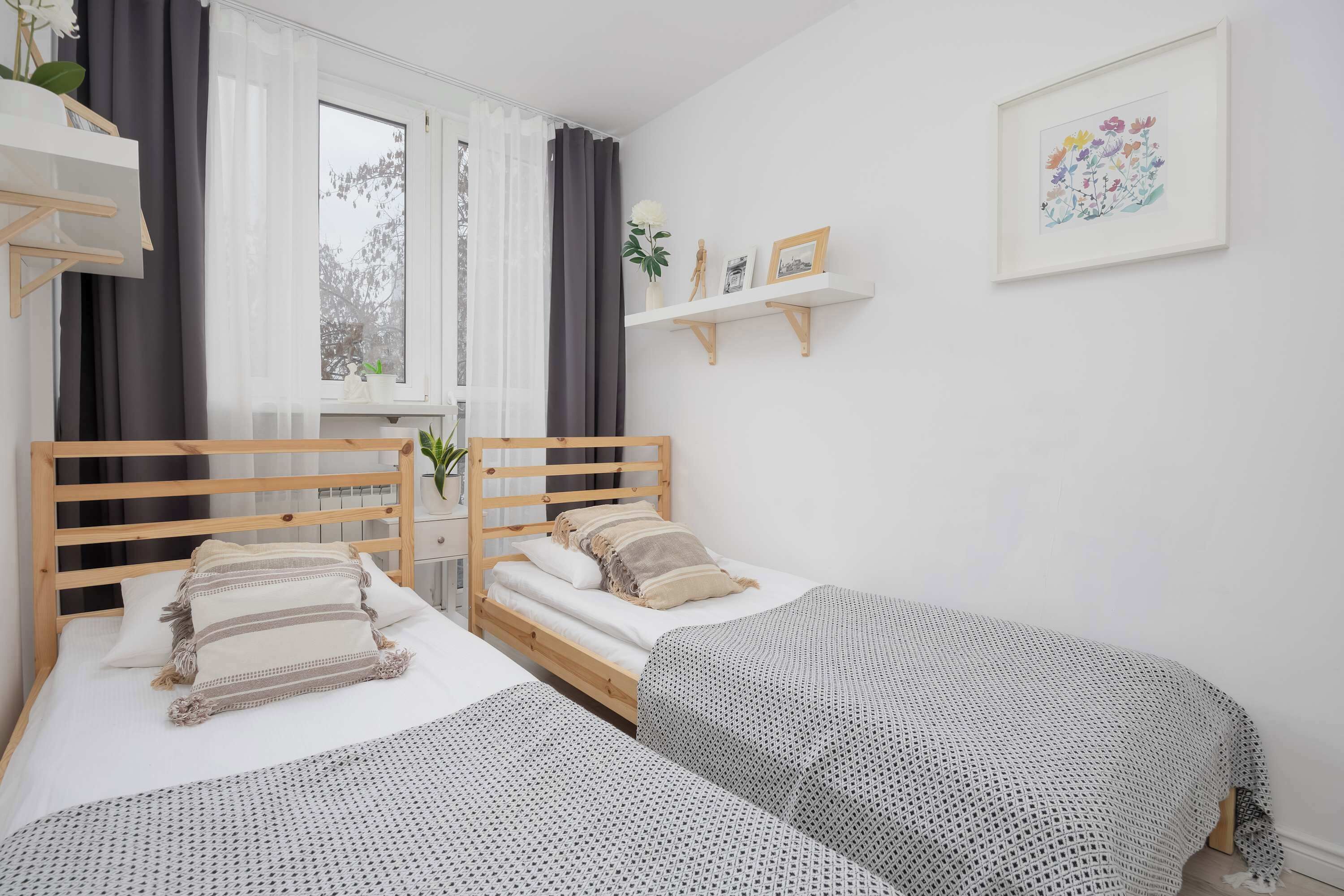 A bedroom with two beds, a window with curtains, a shelf with pictures, and a framed artwork on the wall.
