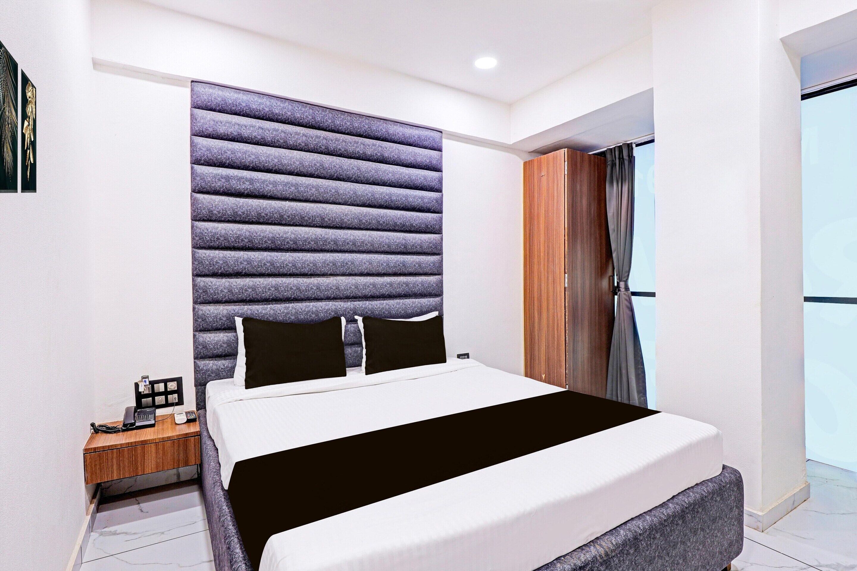 A modern hotel room with a large bed, a wooden nightstand, a headboard with a textured design, and a view of the outdoors through a window.