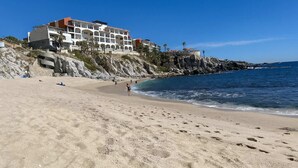 A beach with a hotel on a rocky cliff, clear blue sky, and calm sea. - Marbella Lane - PrimeVilla n Modern Home (Cabo San Lucas)