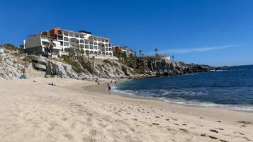 A beach with a hotel on a rocky cliff, clear blue sky, and calm sea.