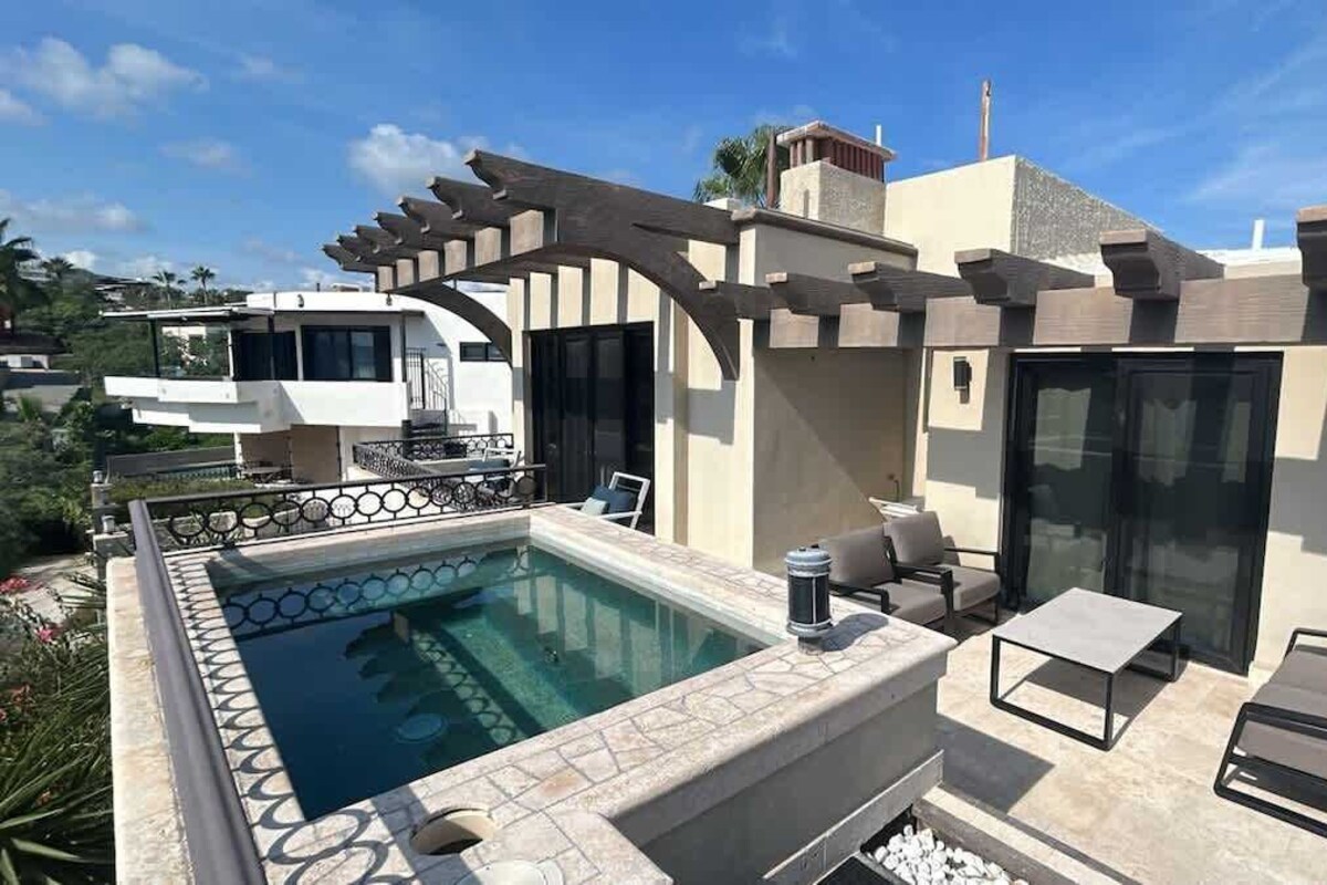 A rooftop pool area with a pergola, outdoor seating, and a view of neighboring buildings.