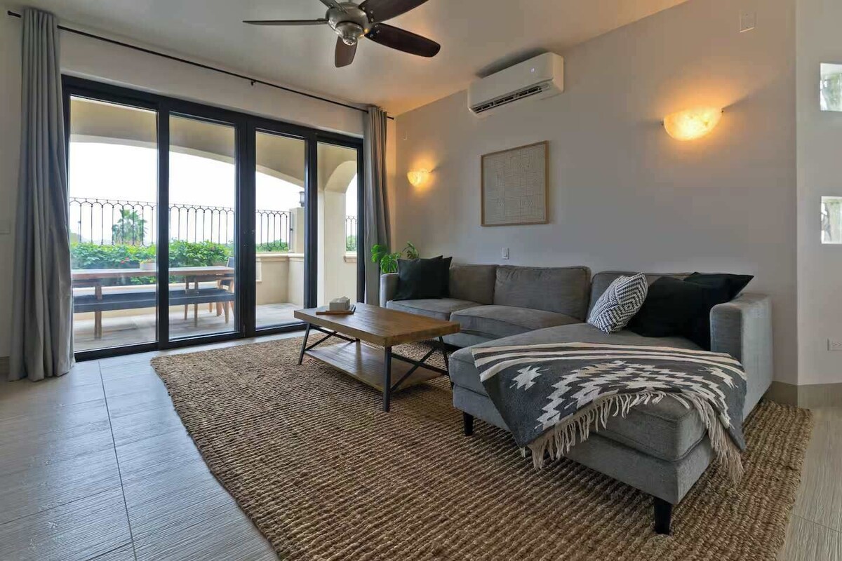 A modern living room with a grey sofa, a wooden coffee table, and a large sliding glass door leading to an outdoor balcony.