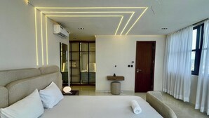 A modern hotel room with a large bed, a bedside table, a mirror, a window with curtains, and a bathroom visible through a glass door. - Phaluxe Shortlets (Abuja)
